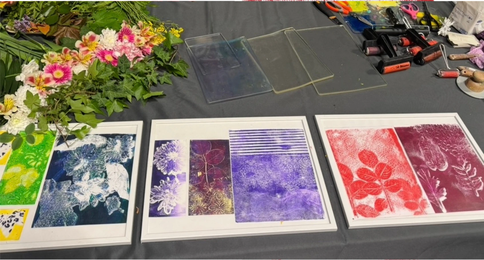 Three framed gel plate monoprints in green, violet and red laid on a studio table beside fresh flowers, clear gel plates and inked brayers.