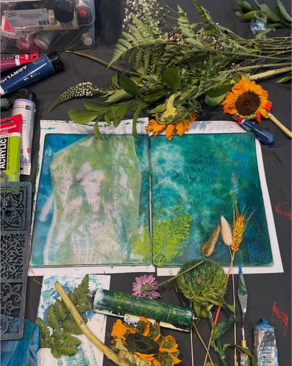 An open art journal printed in teal and green on a black studio table, surrounded by ferns, sunflowers, dried grasses and tubes of acrylic paint.