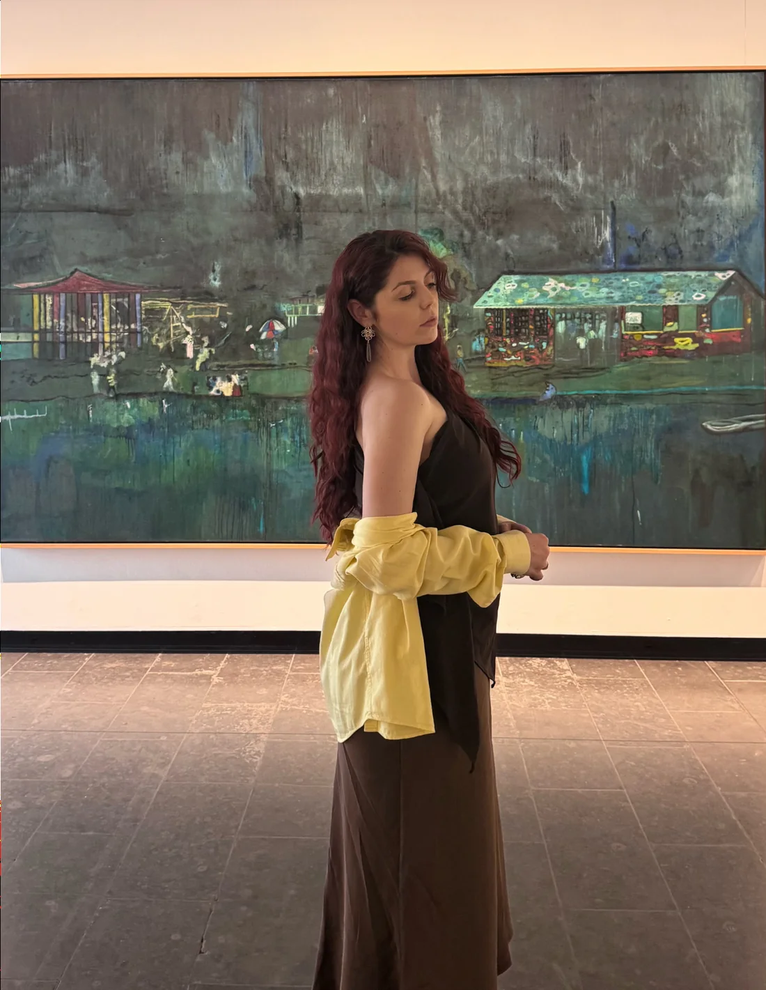 The artist standing in a gallery at the Louisiana Museum of Modern Art, in front of a large landscape painting of huts on dark water.