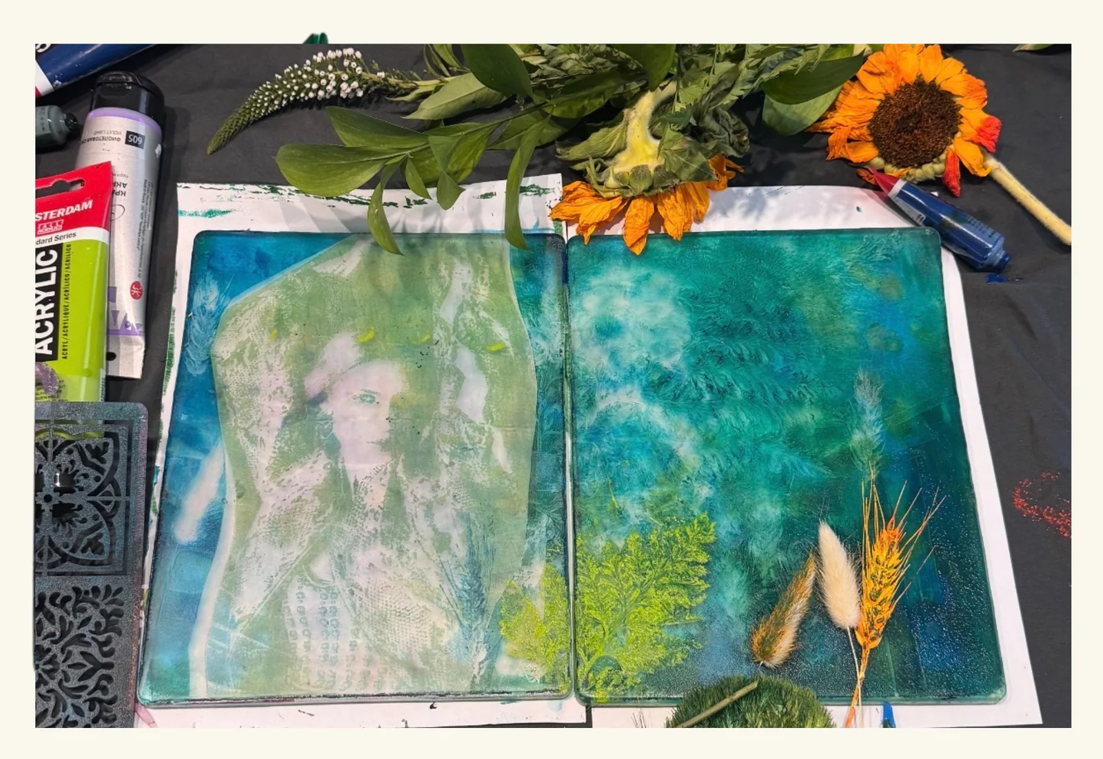 An open art journal spread printed in teal and green - one page holding a ghostly female figure, the other soft fern prints - laid on a black cloth among fresh flowers, a sunflower, dried grasses, a stencil and tubes of acrylic paint.